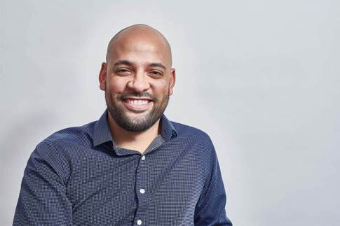 Smiling bald man wearing a blue small-check button-up shirt sits slightly turned toward the camera against a plain light-gray background.