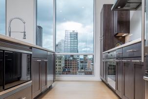 A modern galley kitchen with dark wood cabinets and stainless appliances frames a floor-to-ceiling window overlooking mid-rise city buildings under a cloudy sky.