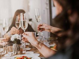 Champagne flutes clink as diners raise glasses around a table set with plates, salad, a white floral centerpiece and glassware in a softly lit restaurant dining setting.