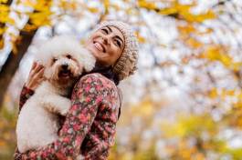 Young woman joyfully holds small fluffy white dog on her shoulder, both smiling amid golden autumn trees and blurred yellow leaves; she wears a knit hat and floral coat.