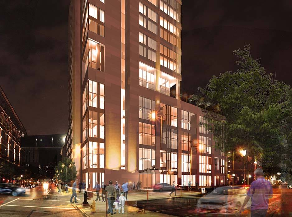 Tall, illuminated glass-and-stone high-rise fills an urban intersection, its warm-lit windows glowing as pedestrians cross and blurred cars pass along a tree-lined nighttime city street.