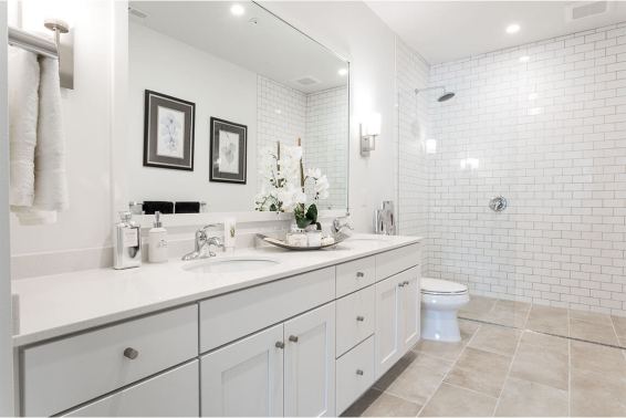 White double-cabinet vanity with countertop sink and decorative orchid sits beneath a large mirror, beside a glass-enclosed subway-tile shower and toilet on a light tiled floor.