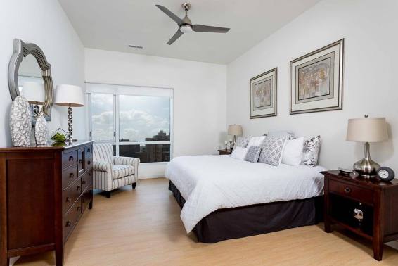 Large bed dressed in white linens anchors a bright, minimalist bedroom; wooden dresser, matching nightstands and striped armchair sit by a window, ceiling fan overhead and city view outside.