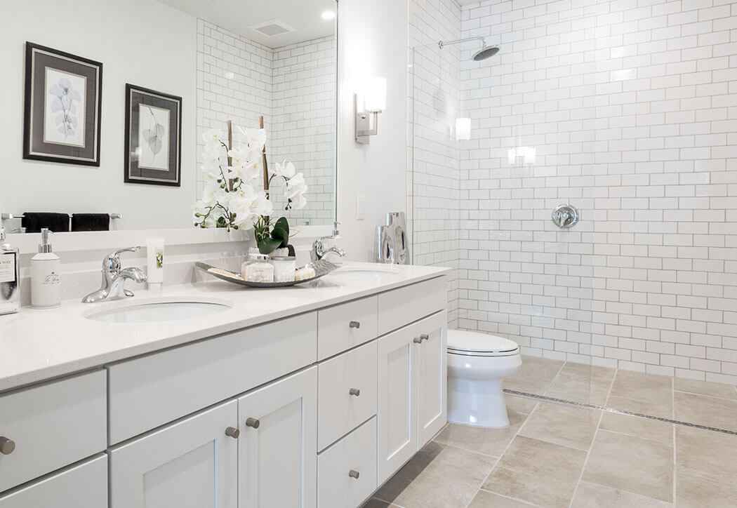 White double-sink vanity displays a tray with orchids and toiletries, set against a mirror in a bright bathroom with subway-tiled glass shower and toilet on beige tile.
