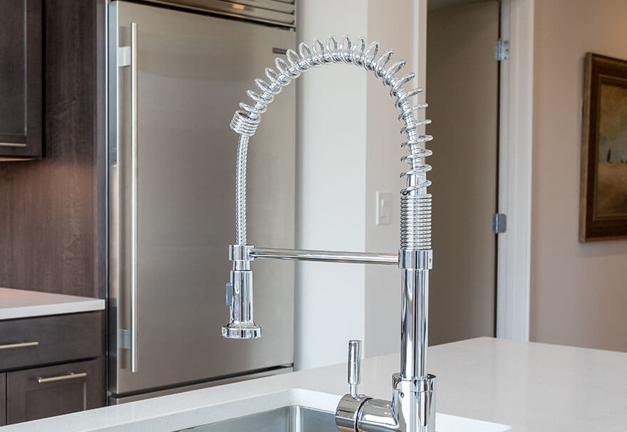 Chrome spring-coil pull-down kitchen faucet extends over a stainless sink, stationary, mounted on a white quartz island countertop