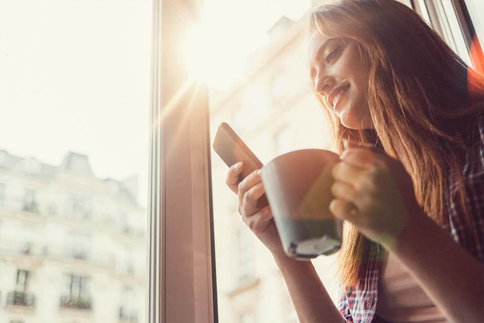 Young woman smiles while holding a smartphone and coffee mug, looking out a sunlit window overlooking blurred city buildings.