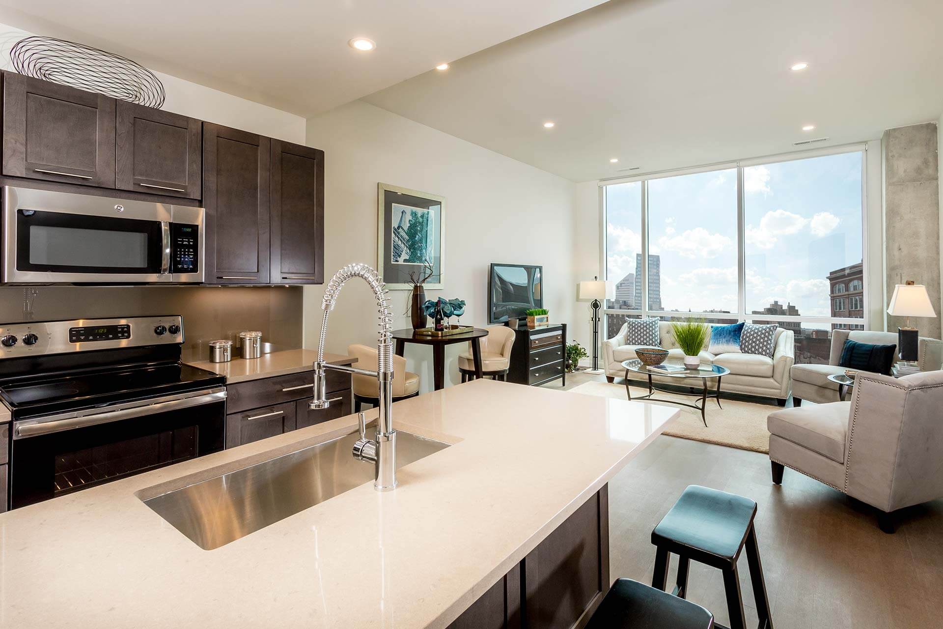 Quartz kitchen island hosts a tall coil-spring faucet, separating dark cabinetry and stainless appliances from a sunlit living room with sofa, glass coffee table, floor-to-ceiling windows and city view.