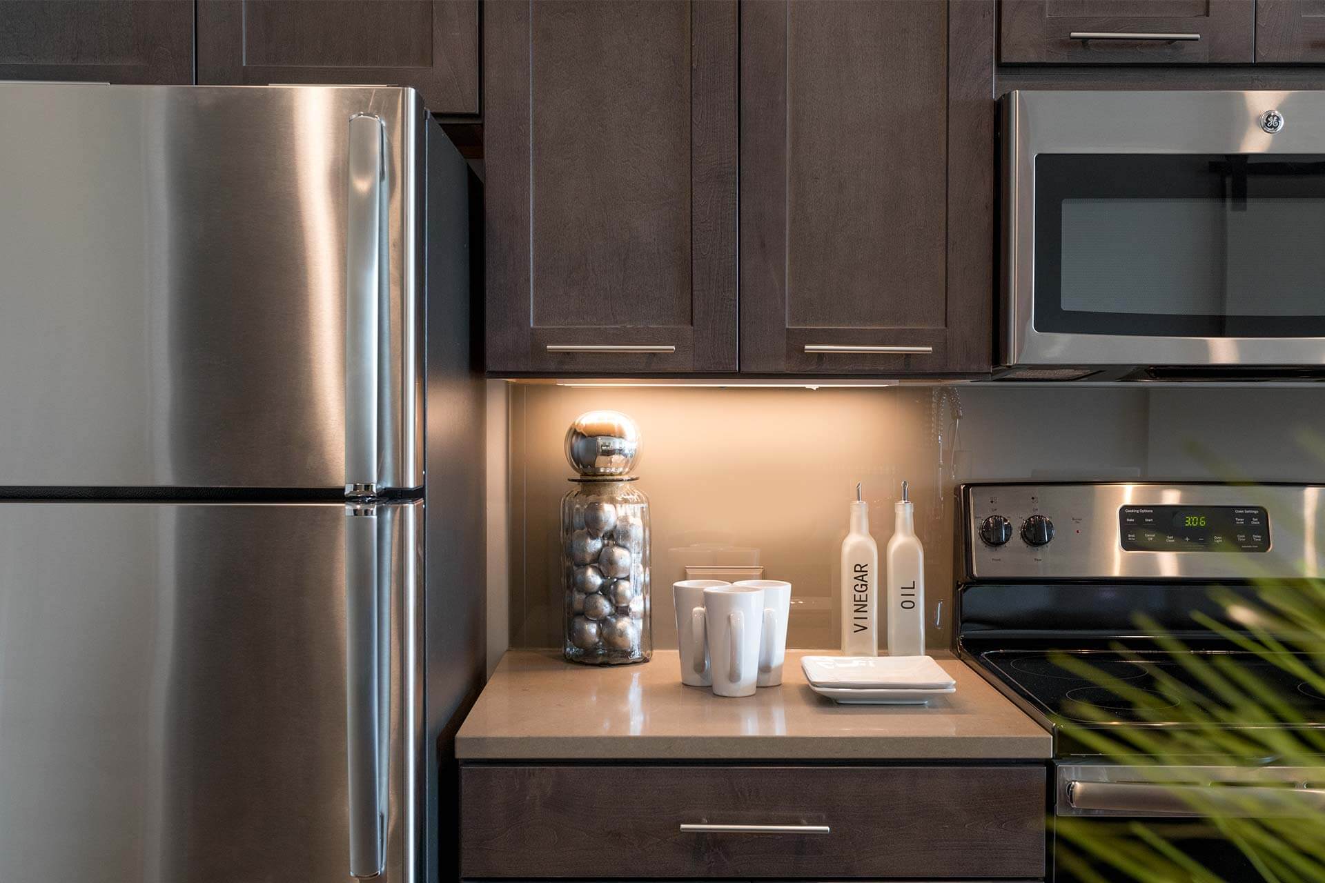 A glass jar filled with metallic spheres and two bottles sit on a countertop beneath warm under-cabinet lighting, between stainless appliances and dark wood cabinets.