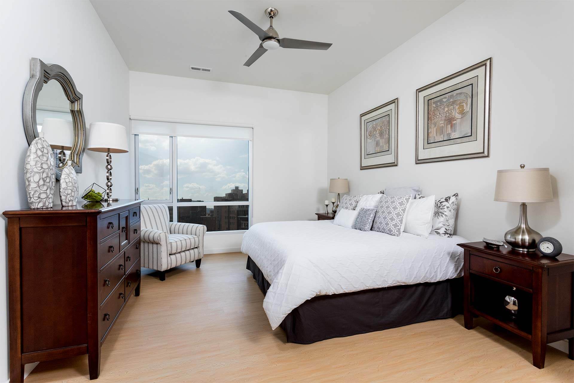 A white king-size bed rests against a light wall, flanked by dark wood nightstands with lamps