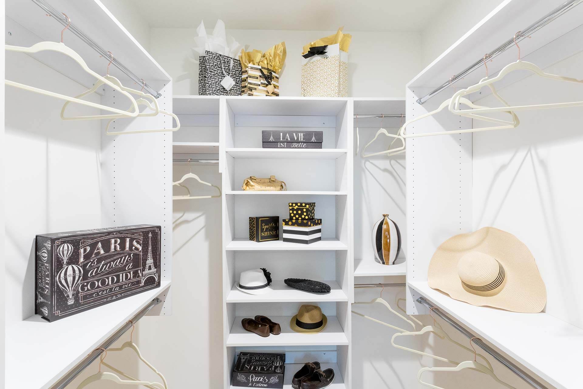 Center shelving unit stores hats, shoes, decorative boxes and gift bags, while empty hangers hang from rods in a bright white walk-in closet.