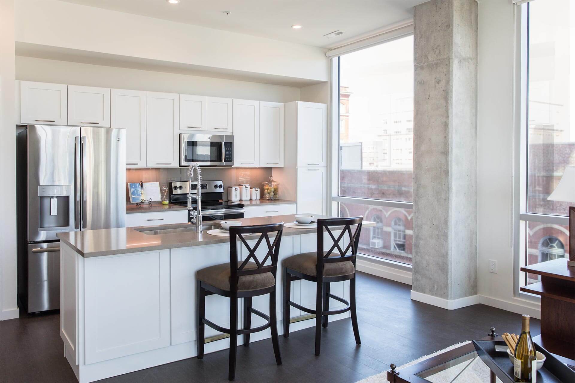 Kitchen island holds two dark-wood bar stools and place settings, faces stainless appliances with tall faucet, in a bright loft kitchen with floor-to-ceiling windows and concrete column.