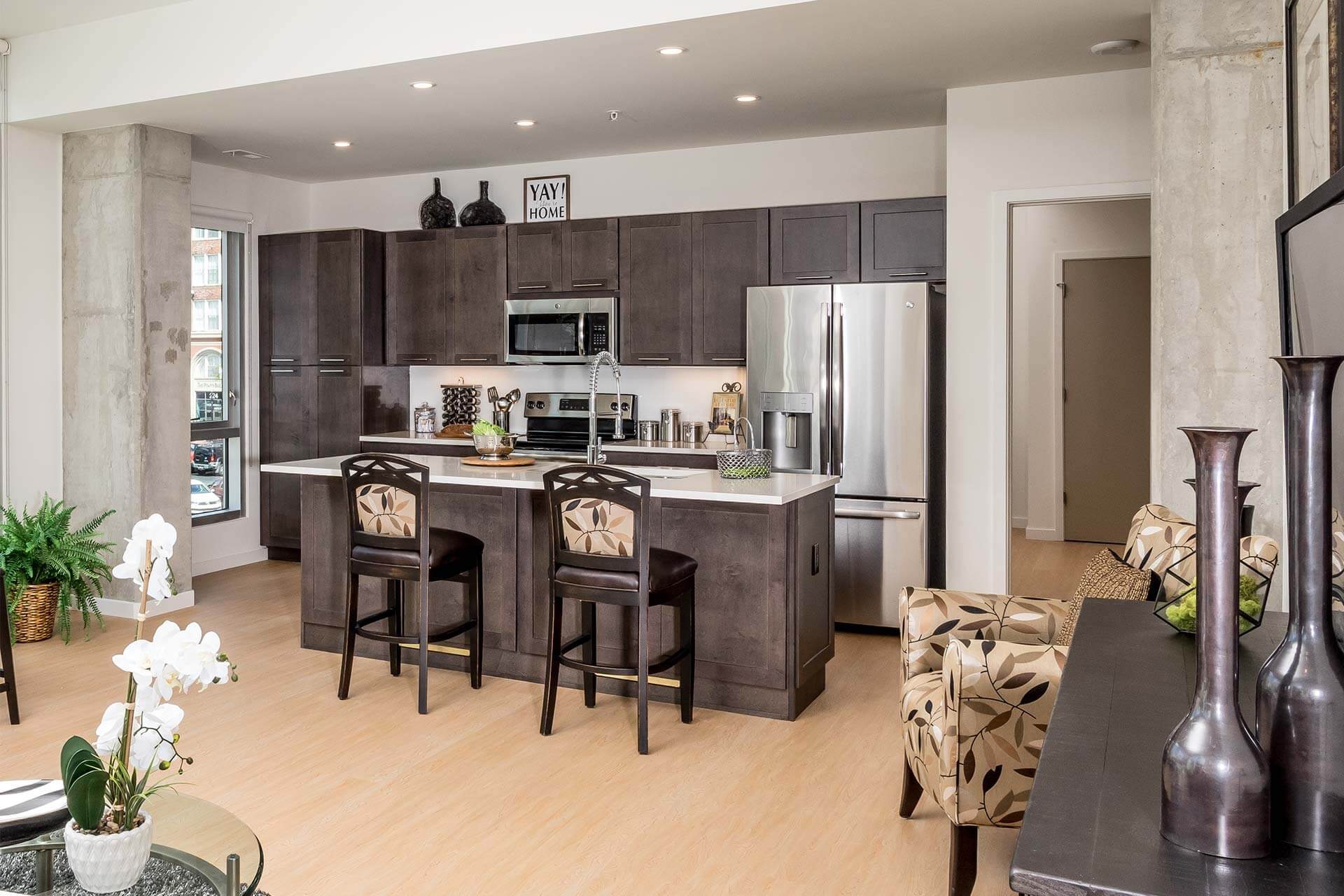 Kitchen island with two patterned bar stools anchors an open-plan living area; island holds sink, faucet and decor, backed by dark cabinets, stainless appliances