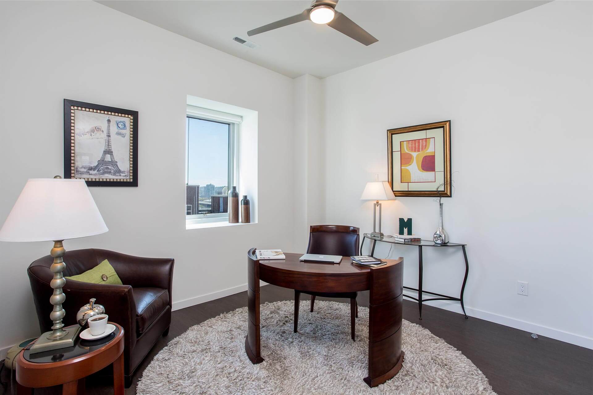 Desk holds a laptop and books on a round rug in a bright office with a leather armchair, a side-table lamp, framed artwork, and a small window showing distant buildings.