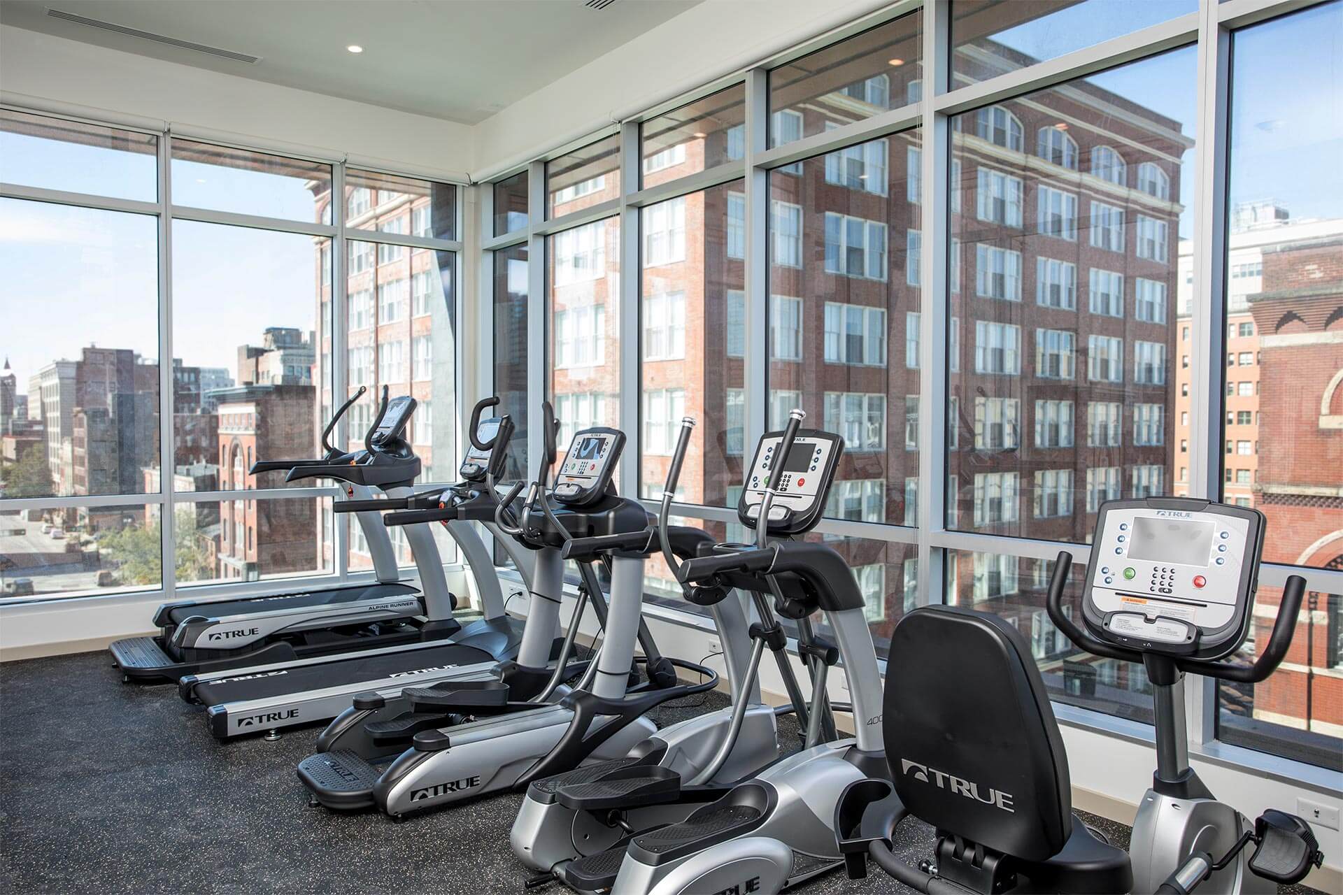 Exercise machines sit unused facing floor-to-ceiling windows, overlooking adjacent red-brick apartment and office buildings in a sunlit urban gym.