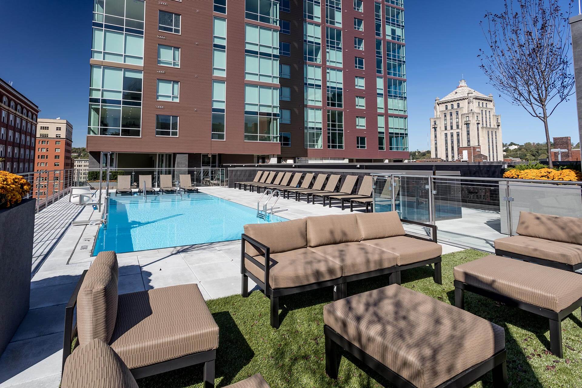 Rooftop pool sparkles, framed by rows of sun loungers and cushioned seating on green turf, set against a modern glass-and-brick apartment tower and a distant historic high-rise.