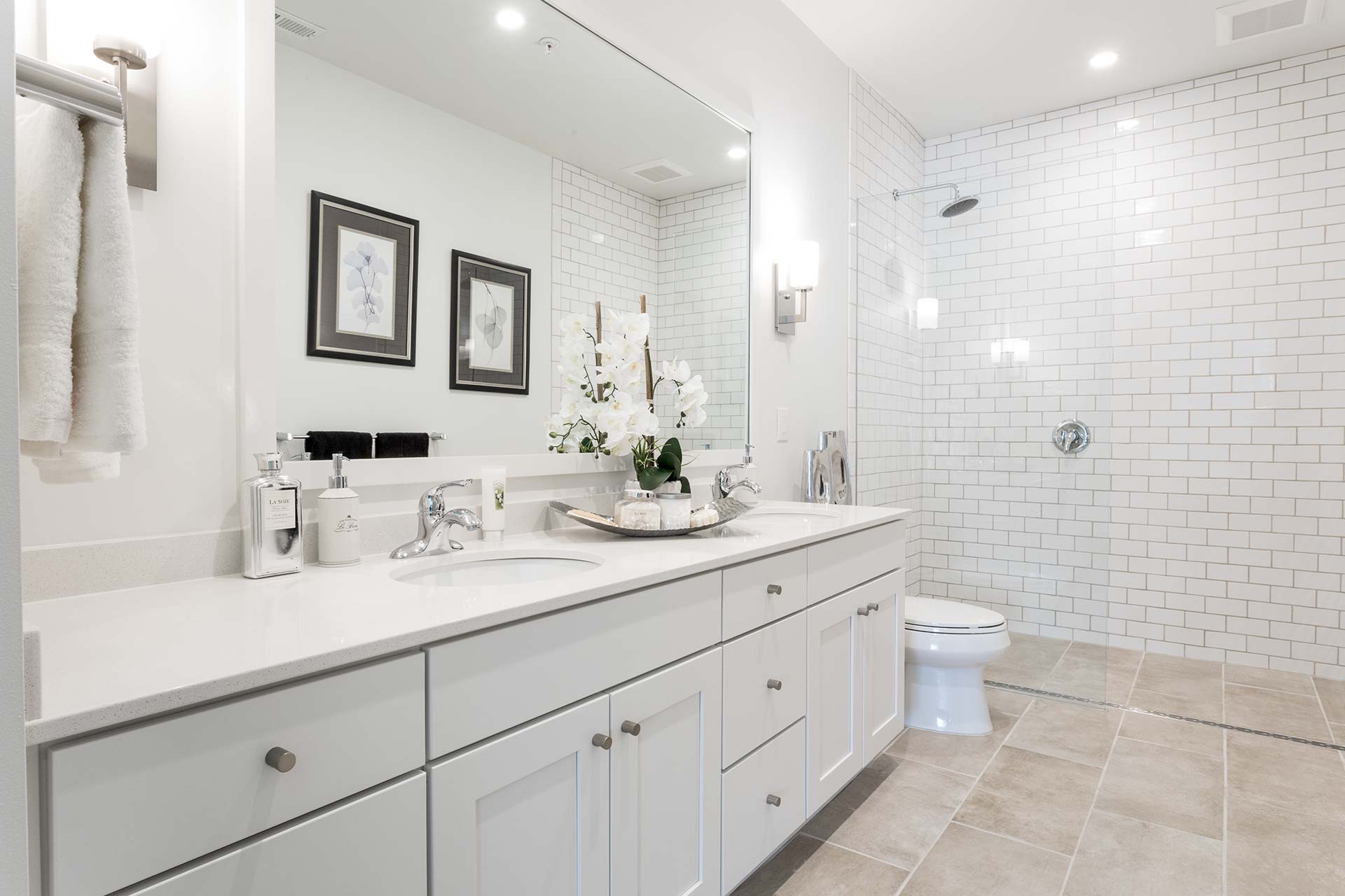 A white double-sink vanity displays polished chrome faucets and a tray of toiletries and orchids, reflecting in a large mirror; a glassless tiled shower and toilet fill the bright bathroom.
