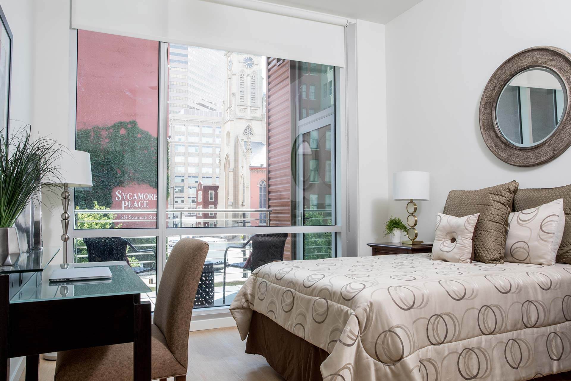Bed dressed with circular-patterned comforter and decorative pillows sits beside a desk and lamp, facing floor-to-ceiling window revealing a balcony and city buildings.
