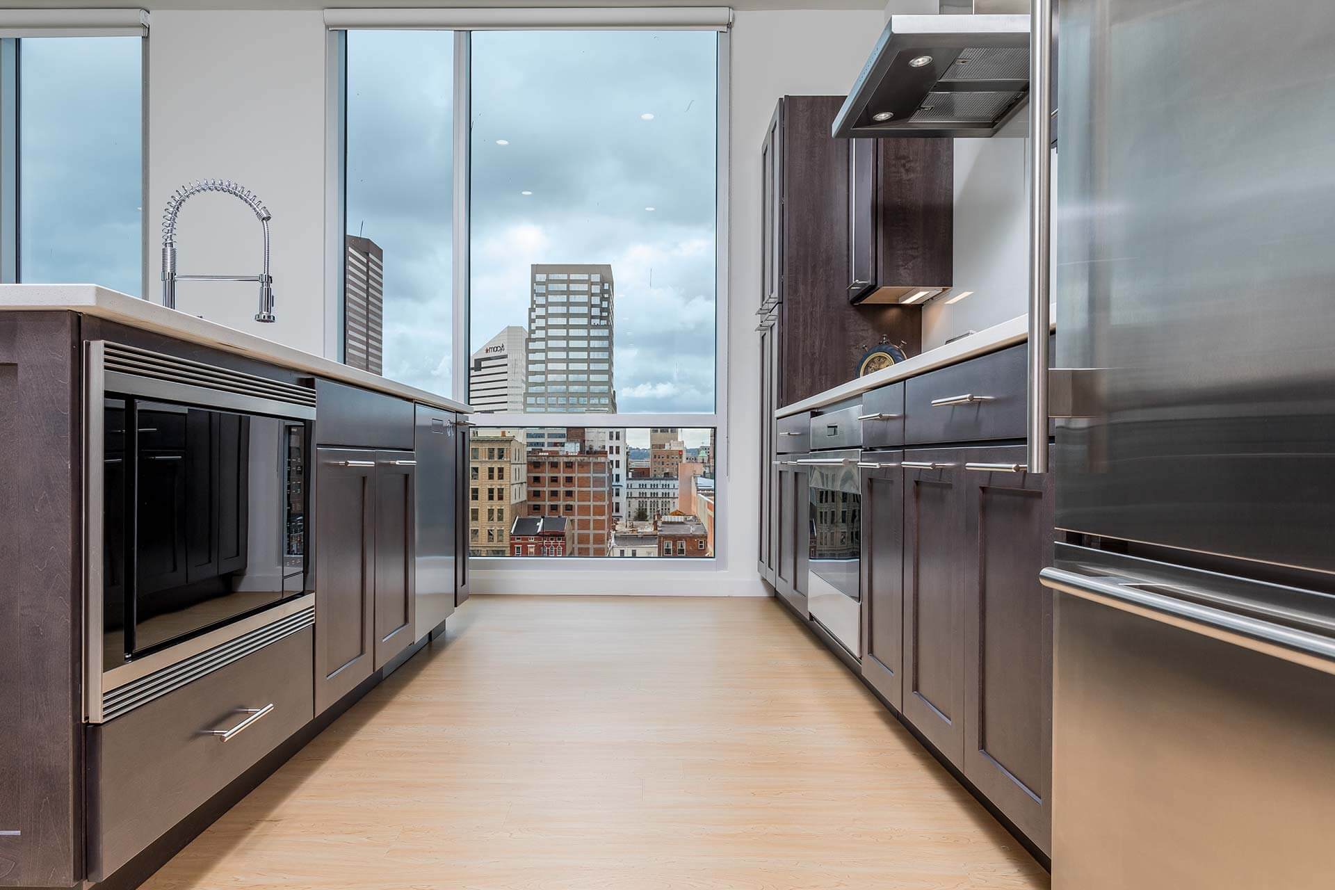 Kitchen island with a tall stainless spring faucet faces a floor-to-ceiling window revealing a cloudy city skyline; dark wood cabinets and stainless appliances flank a light wood floor.
