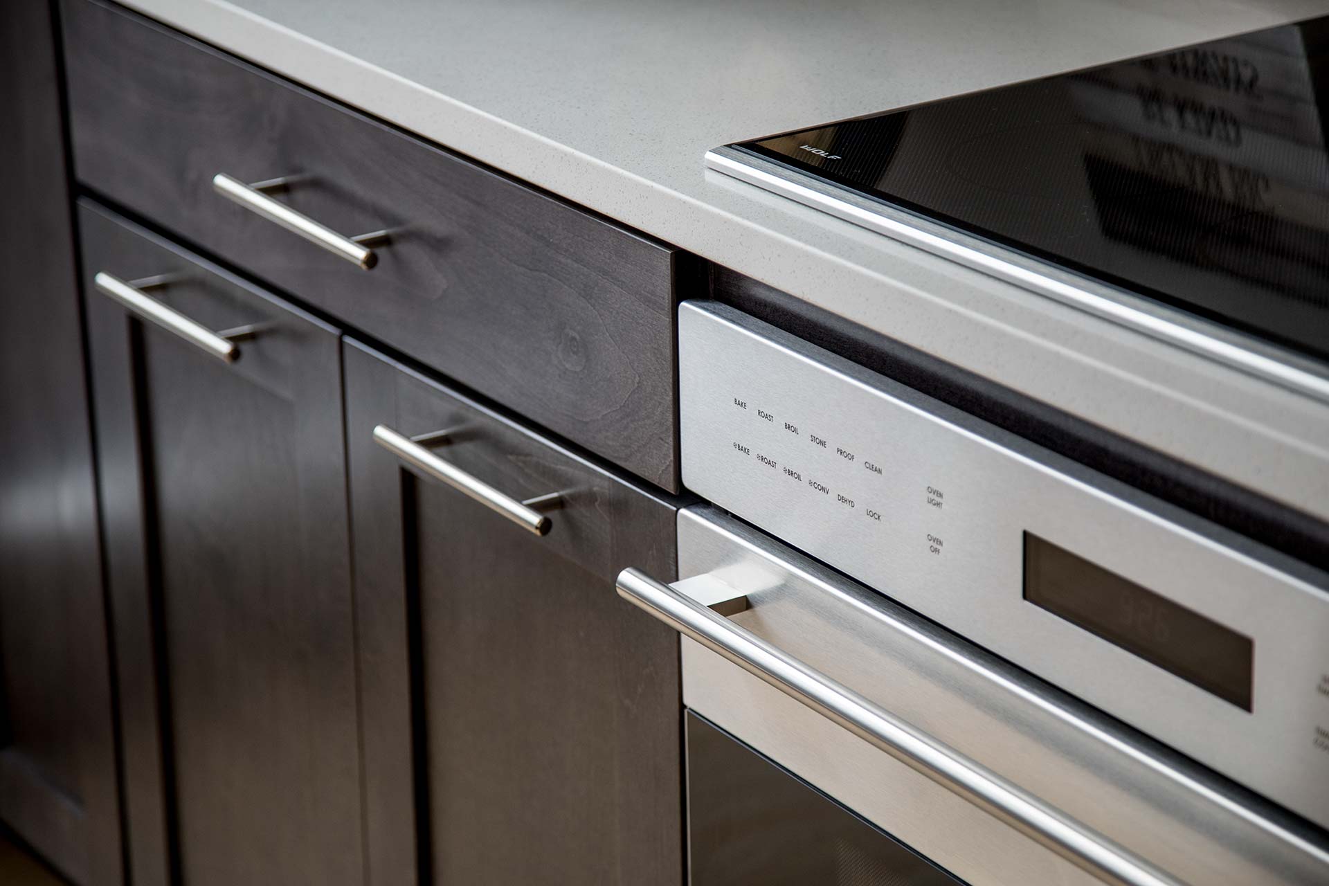 Stainless steel oven front and long bar handle sit flush with dark wood cabinet doors and matching horizontal pulls beneath a light quartz countertop and black glass cooktop.