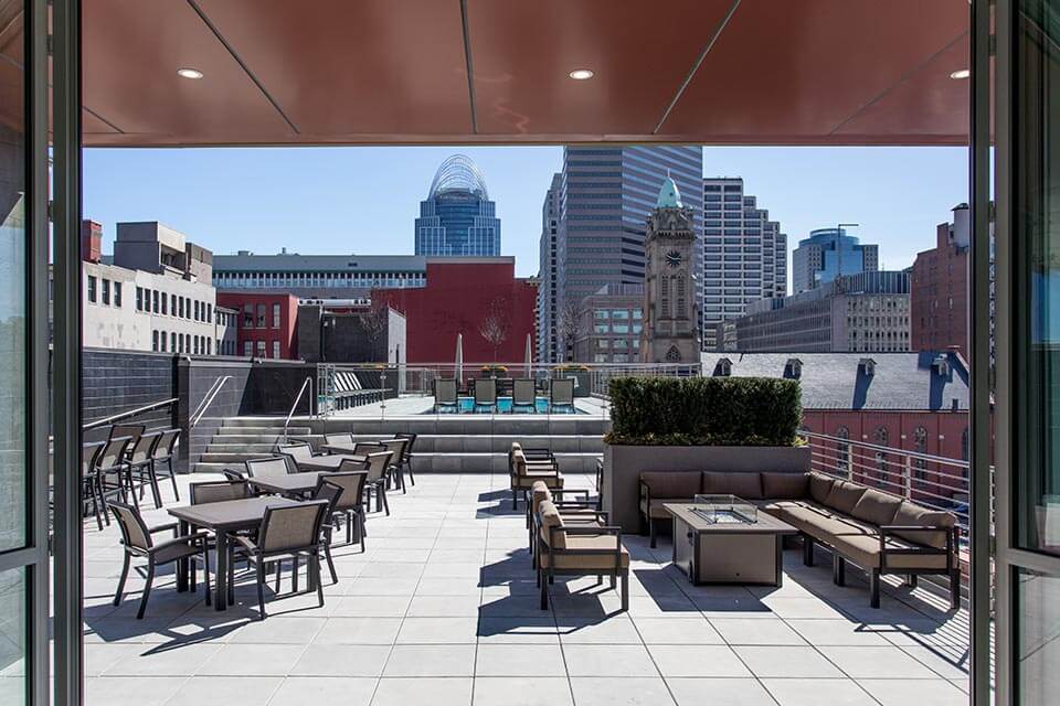 Rooftop terrace with lounge and dining furniture arranged around a low fire table and small pool, overlooking downtown skyscrapers and a clock tower under a clear blue sky.