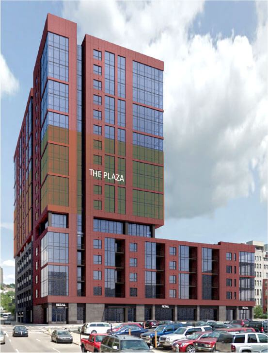 A tall red-brick-and-glass mixed-use tower stands above a low retail podium overlooking a crowded parking lot and urban street beneath a cloudy sky
